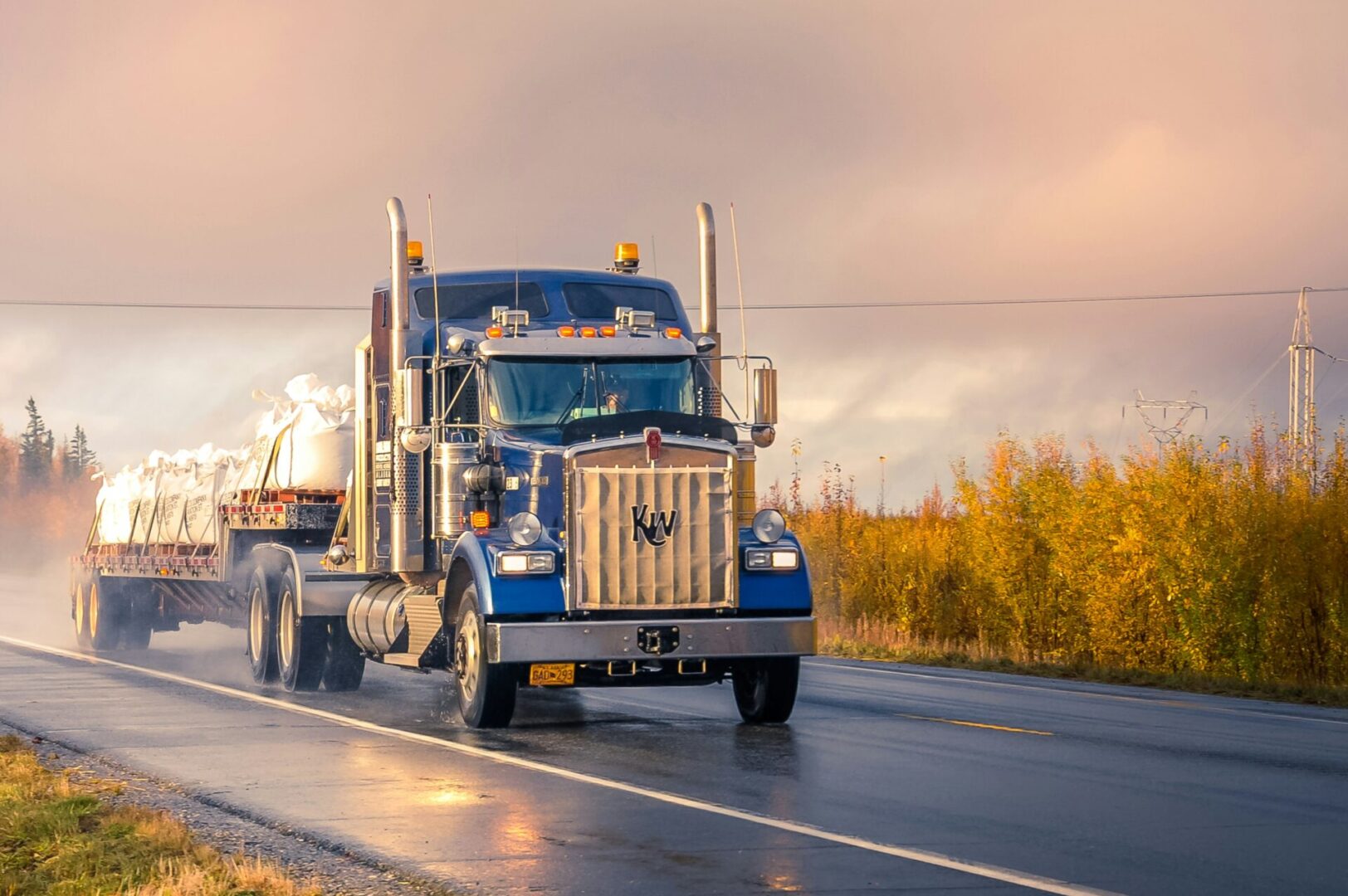 A blue semi-truck driving on a wet road during sunset.